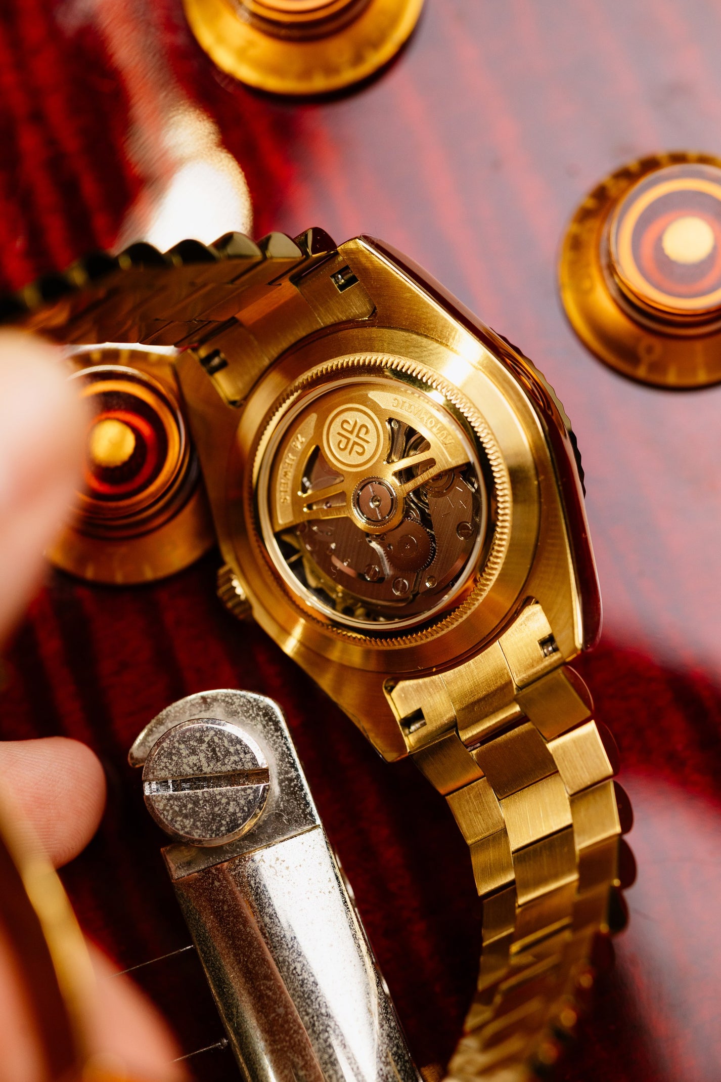 The Just Date, spate transparent din aur cu mecanism automatic vizibil, reglat cu o unealta, pe fundal cu monede aurii.
