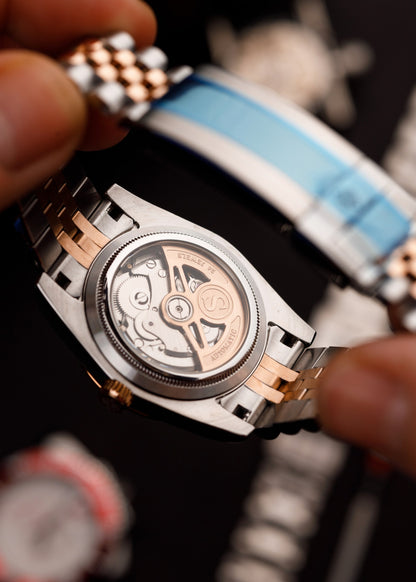 The LadyJust, spate transparent cu mecanism automatic vizibil, model bicolor, lângă o brățară alb-albastră.
